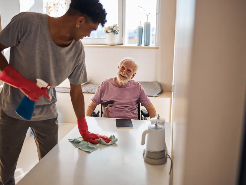 A support worker wiping down the kitchen bench, talking with an older man in a wheelchair