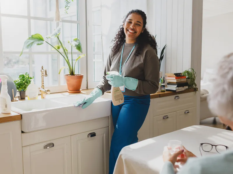A support worker cleaning the kitchen while her client sits nearby with a cup of tea