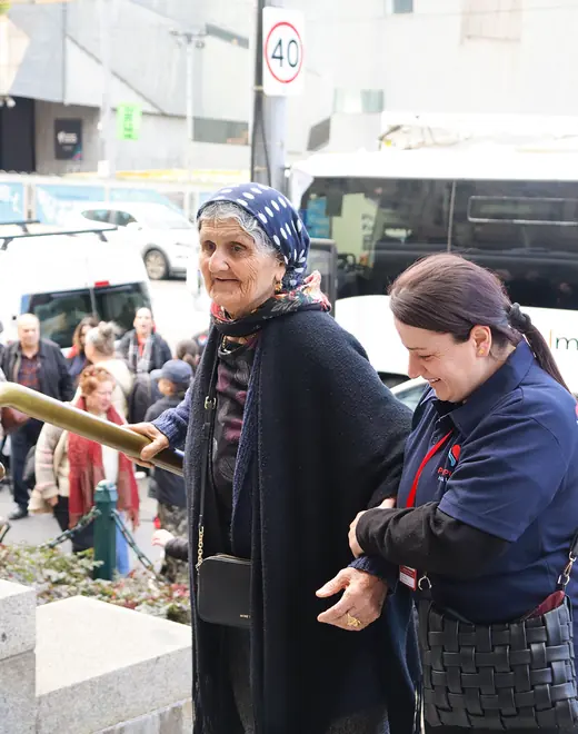A support worker steadying an older woman by the arm as she walks up a step outdoors