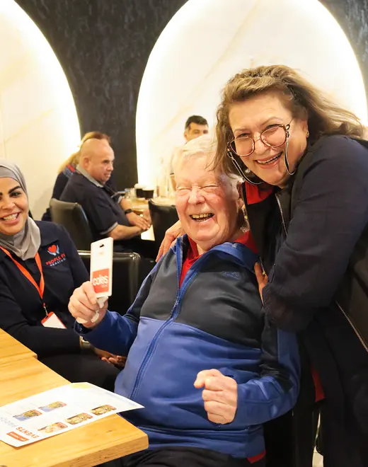 An older man laughing as he holds up a prize card, with a People First Healthcare carer beside him