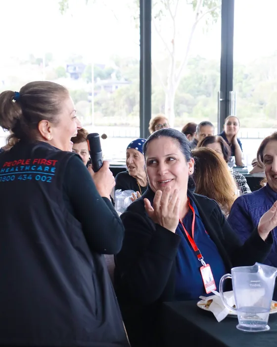 A People First Healthcare staff member with a microphone at a community lunch, carers and clients applauding
