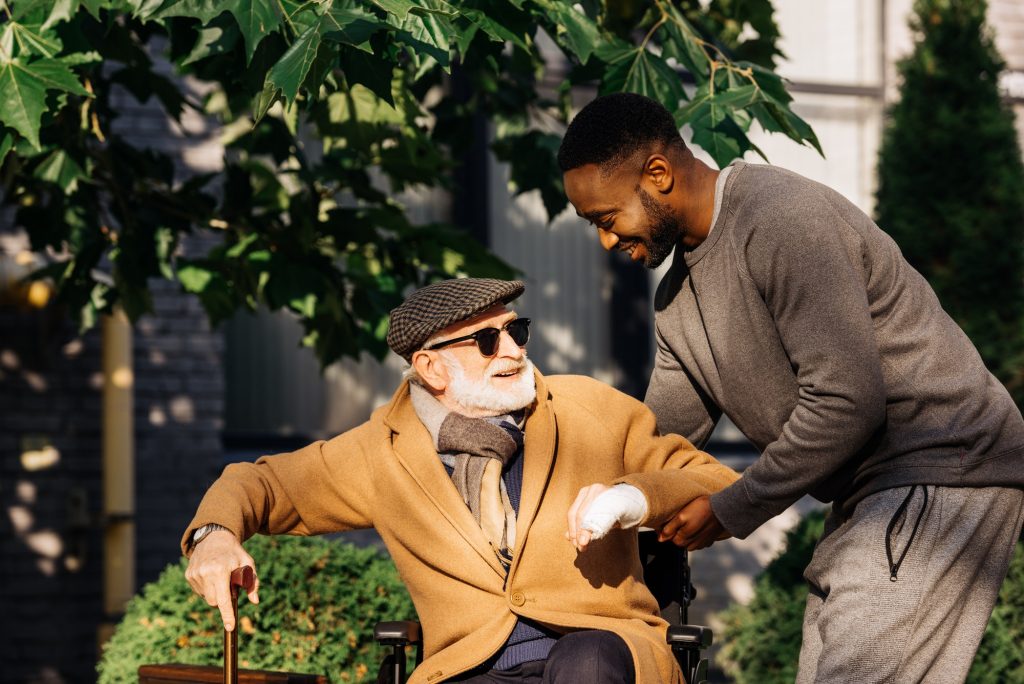 happy african american man helping senior disabled man to get up from wheelchair on street